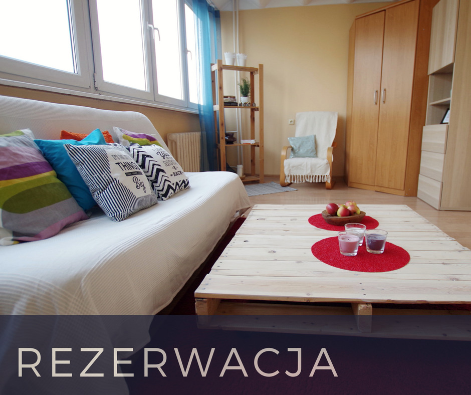 Jasne wnętrze pokoju z sofą z kolorowymi poduszkami, stolikiem kawowym z palet i drewnianymi meblami, z widocznym napisem 'Rezerwacja'.