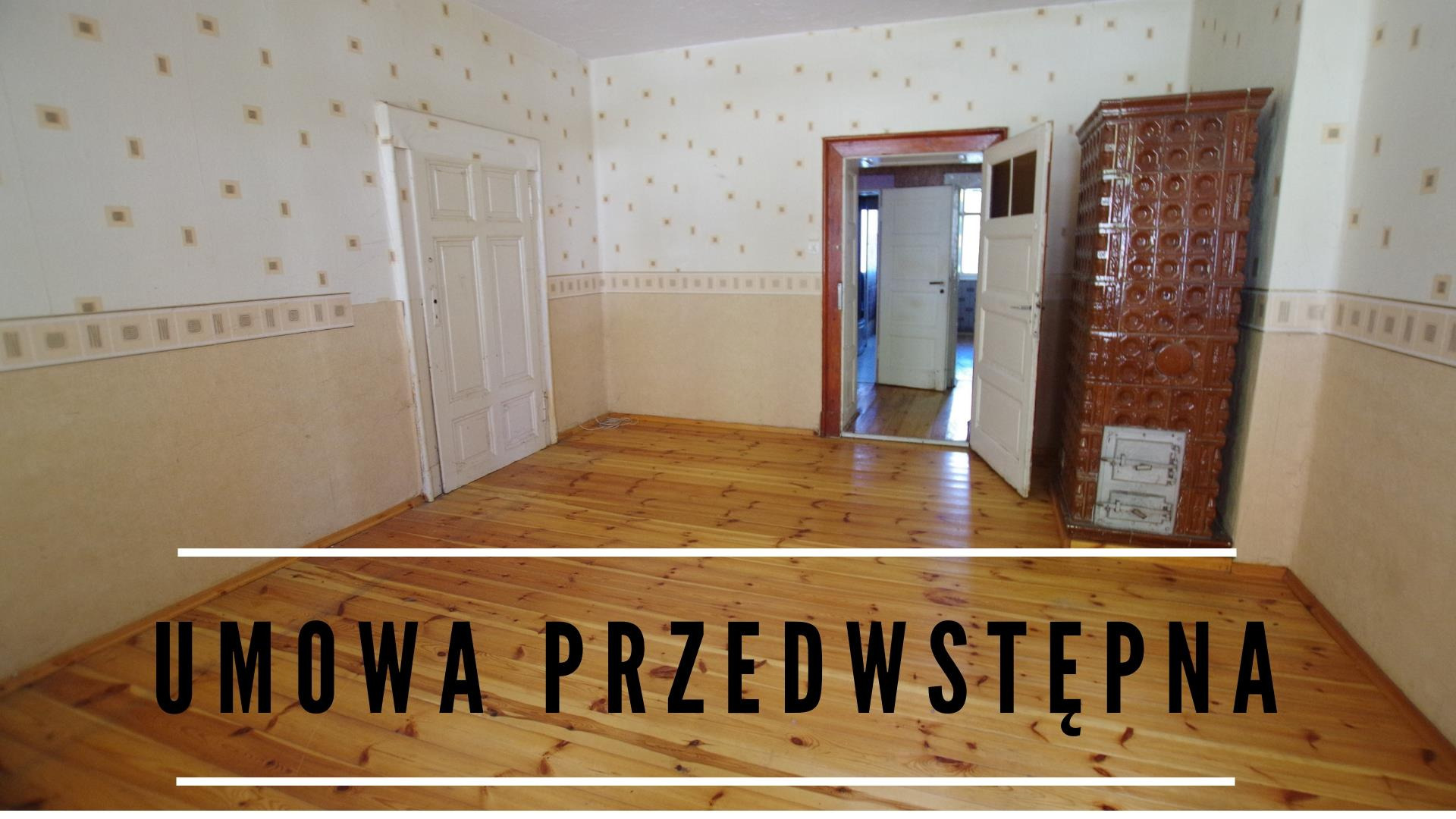 Pusty pokój z drewnianą podłogą, piecem kaflowym w rogu i dwoma parami drzwi, ściany z tapetą w drobne wzory.