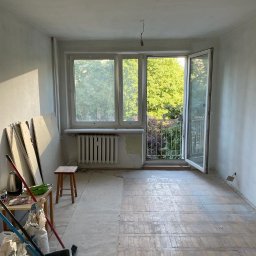 Erwin- remonty - Pokój w trakcie remontu: surowe ściany, częściowo zakryta podłoga, otwarte drzwi balkonowe z widokiem na zieleń, grzejnik pod oknem i akcesoria malarskie