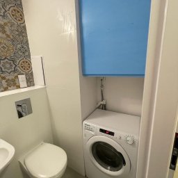 Erwin- remonty - Kompaktowa łazienka z wiszącą toaletą, umywalką i pralką Candy, ukrytą pod niebieską zabudową, na tle płytek w geometryczne wzory.