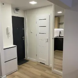 Nowoczesne wnętrze mieszkania po remoncie: widok na przedpokój z szafką na buty, czarne drzwi wejściowe, białe drzwi wewnętrzne z poziomymi frezami i fragment kuchni z czarnymi szafkami...