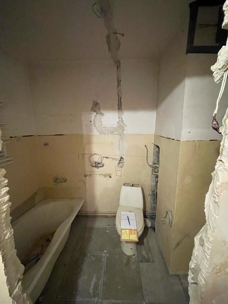 Łazienka w trakcie remontu: widoczne ślady po usuniętych płytkach, odkryte instalacje, zabezpieczona toaleta z kartką na desce, ściany częściowo przygotowane do dalszych prac.
