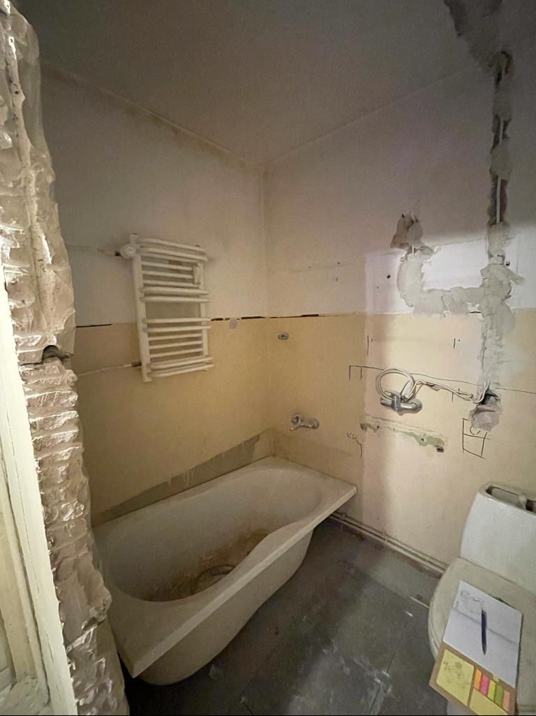Łazienka w trakcie remontu: obite ściany, widoczne instalacje, stara wanna, grzejnik drabinkowy i toaleta z notatkami na desce, ukazujące postęp prac.
