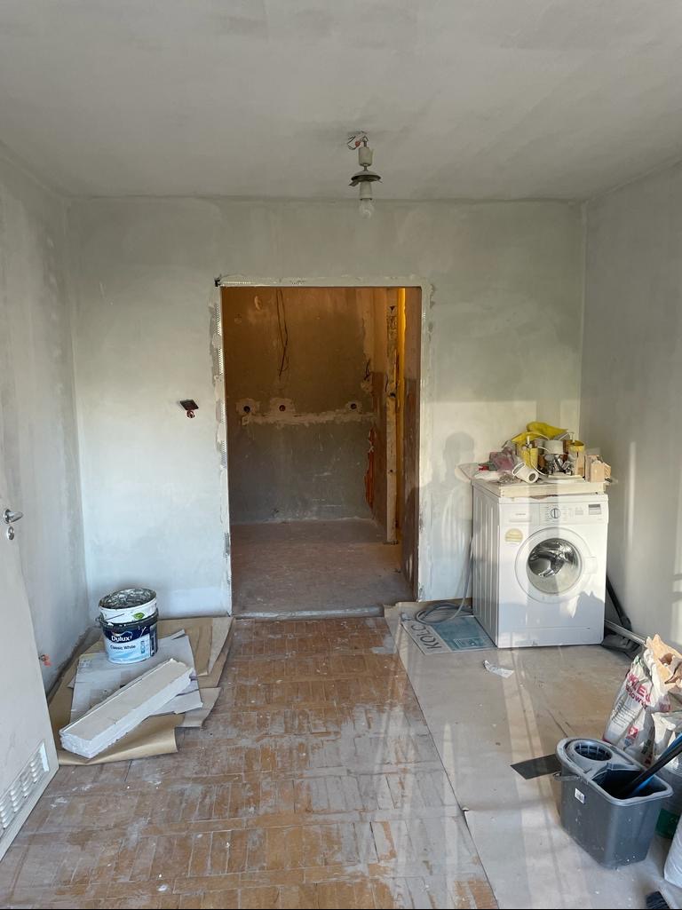 Wnętrze w trakcie remontu: ściany po nałożeniu gładzi, widoczne przygotowania do dalszych prac, podłoga zabezpieczona kartonem, w tle pralka i wiadro z farbą.