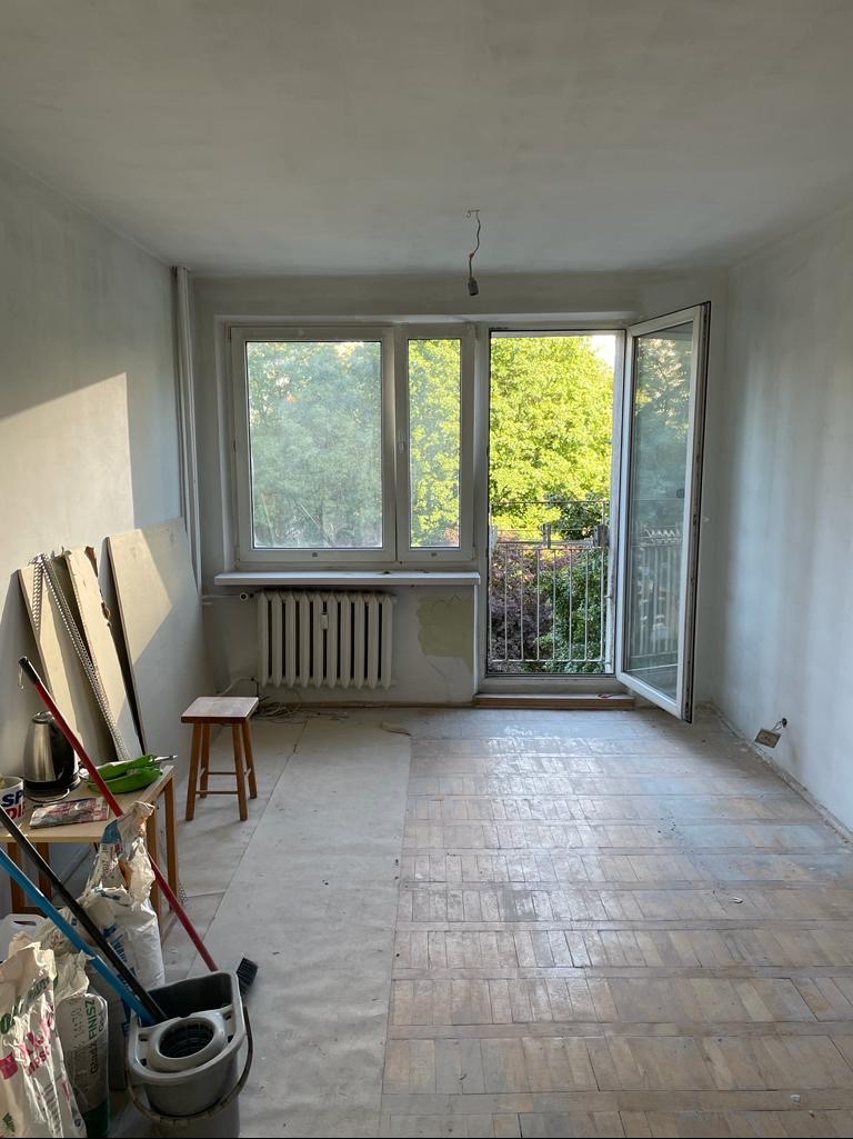 Pokój w trakcie remontu: surowe ściany, częściowo zakryta podłoga, otwarte drzwi balkonowe z widokiem na zieleń, grzejnik pod oknem i akcesoria malarskie