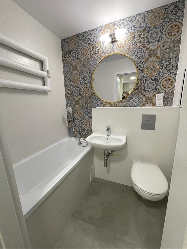 Nowoczesna łazienka z wanną, umywalką i toaletą, wykończona płytkami w marokański wzór na ścianie za okrągłym lustrem.