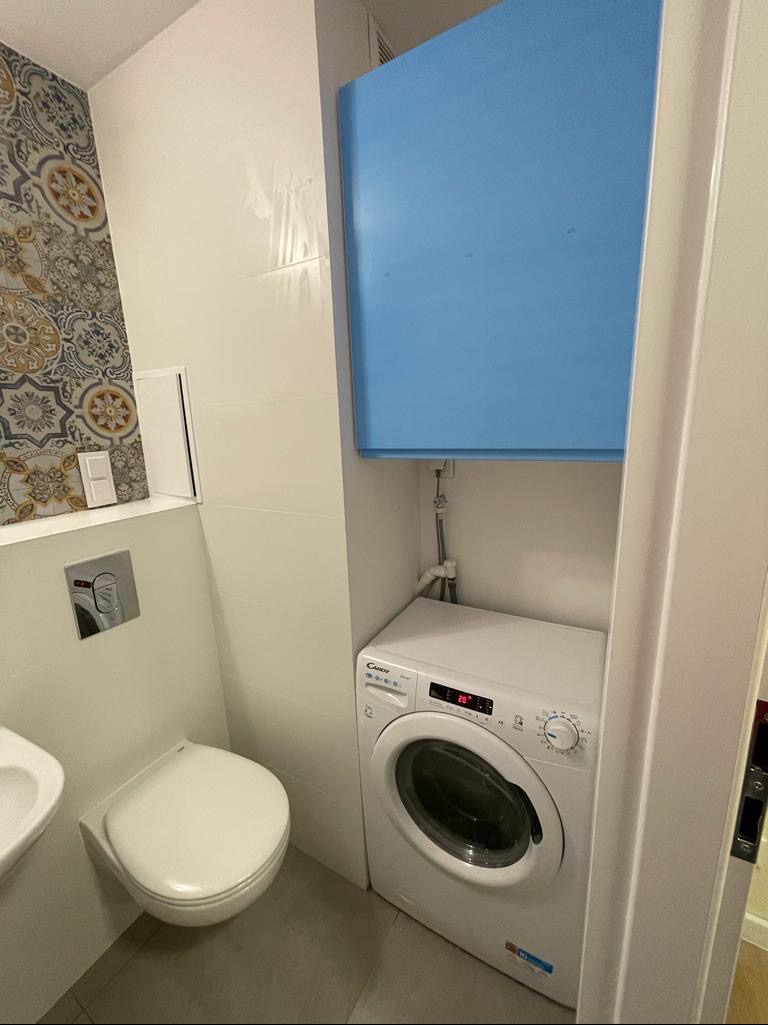 Kompaktowa łazienka z wiszącą toaletą, umywalką i pralką Candy, ukrytą pod niebieską zabudową, na tle płytek w geometryczne wzory.