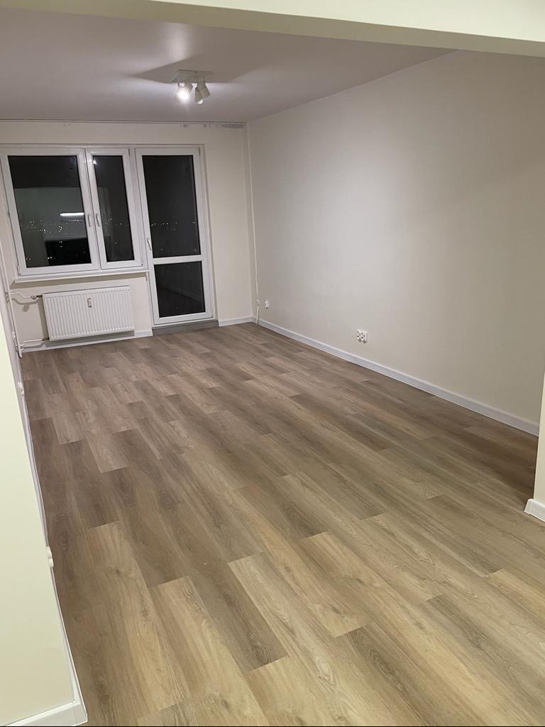 Puste, jasne pomieszczenie z nową podłogą laminowaną imitującą drewno, widoczne okno balkonowe z grzejnikiem pod spodem, surowe ściany przygotowane pod malowanie.