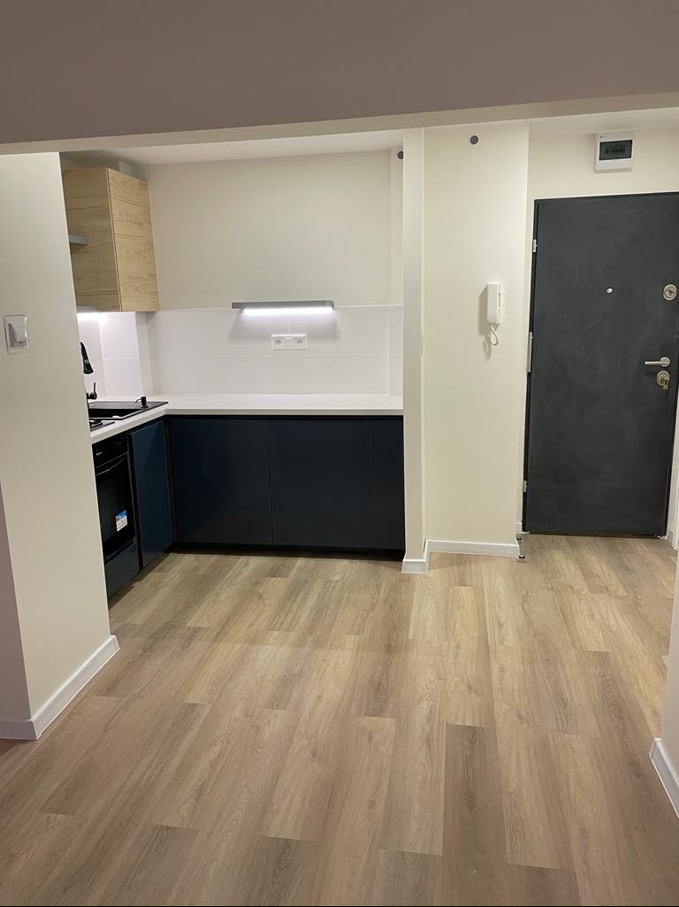 Widok na wykończone wnętrze mieszkania: ciemnoniebieskie szafki kuchenne z białym blatem, drewnopodobna podłoga, białe ściany i czarne drzwi wejściowe.