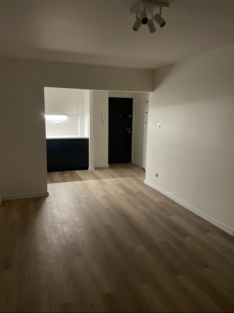 Wnętrze wykończonego mieszkania z panelami podłogowymi w odcieniach brązu, widok na kuchnię z zabudową w kolorze granatowym oraz białe ściany i sufit z nowoczesnym oświetleniem punktowym.