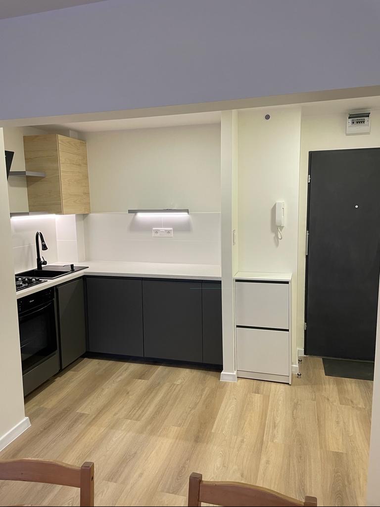 Nowoczesna kuchnia z ciemnymi szafkami dolnymi, jasnym blatem i drewnopodobną podłogą, widok na czarny zlew i płytę grzewczą, oraz białą szafkę na buty przy czarnych drzwiach wejściowych.