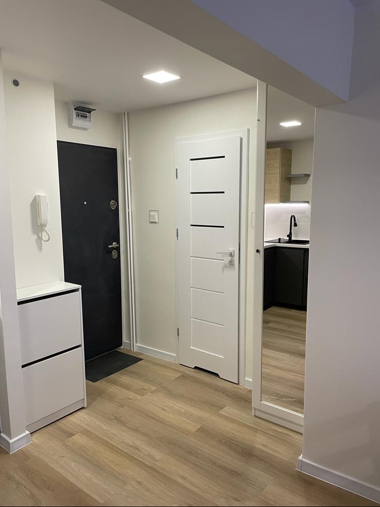 Nowoczesne wnętrze mieszkania po remoncie: widok na przedpokój z szafką na buty, czarne drzwi wejściowe, białe drzwi wewnętrzne z poziomymi frezami i fragment kuchni z czarnymi szafkami...
