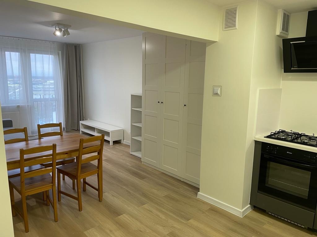 Jasne wnętrze mieszkania z drewnianym stołem i krzesłami, białą zabudową szaf i aneksem kuchennym z czarną płytą grzewczą i okapem.