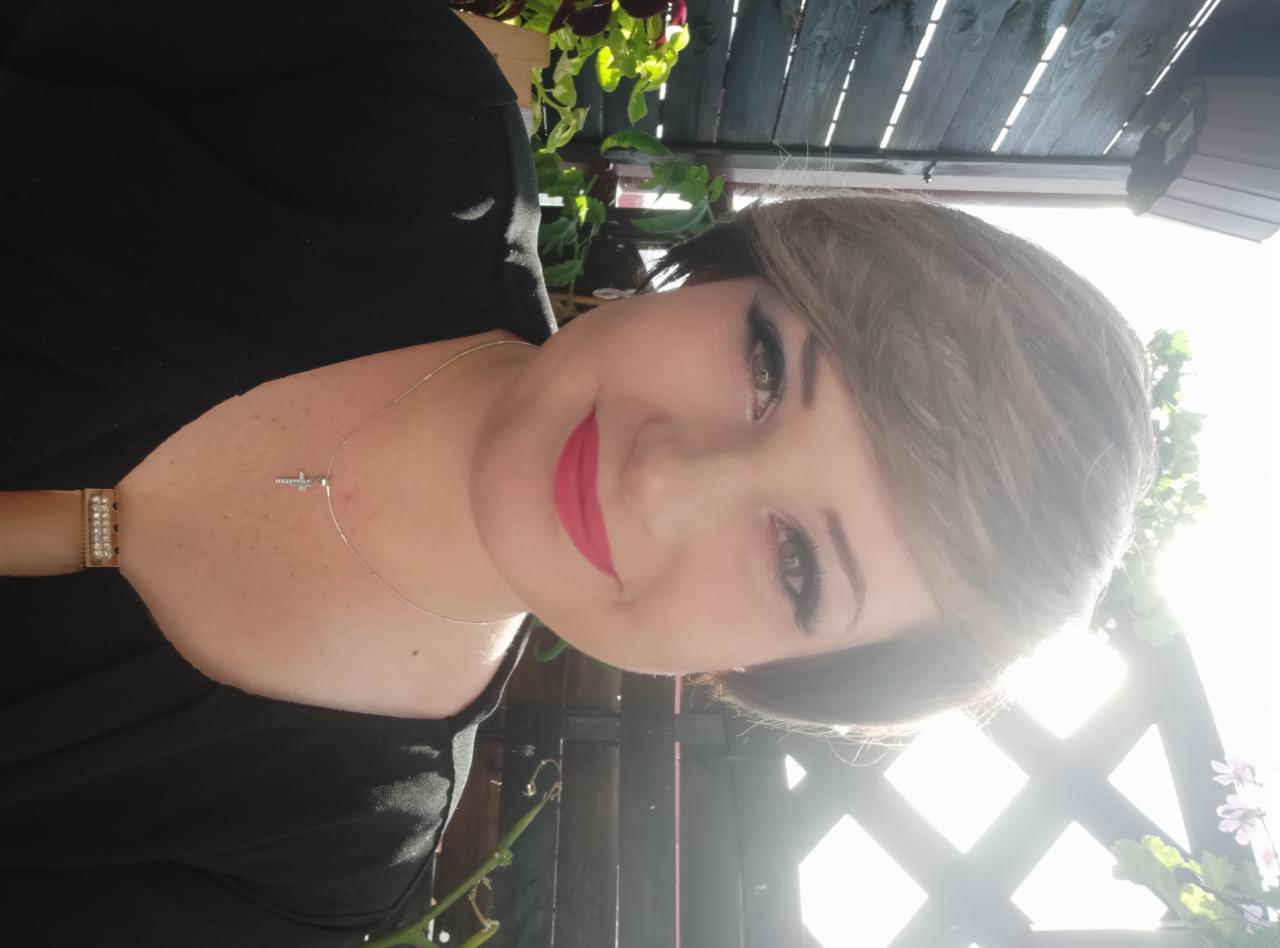 Portret kobiety z krótkimi włosami, w czarnej bluzce, z delikatnym srebrnym naszyjnikiem z krzyżykiem, z mocnym makijażem oczu i ust, na tle drewnianego płotu i zielonych roślin.