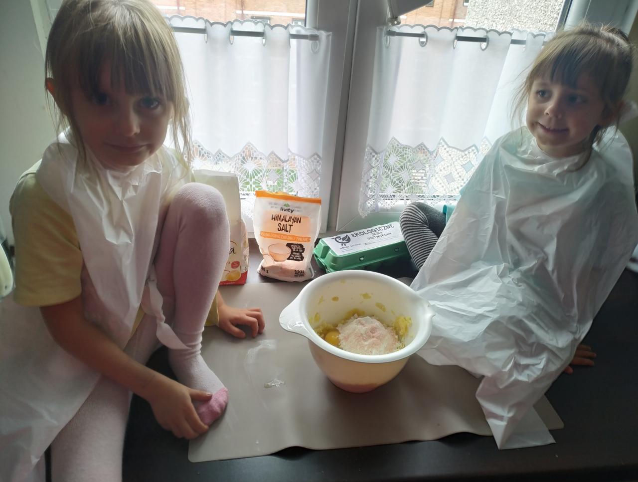 Dwie dziewczynki w plastikowych fartuchach przygotowują posiłek w kuchni, widoczne składniki: mąka, sól himalajska, jajka ekologiczne oraz miska z ziemniakami i mąką.