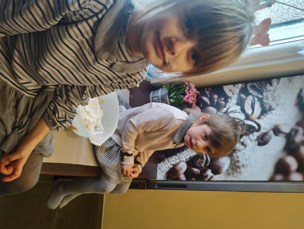 Dwie dziewczynki siedzą przy stole, jedna z plamką mąki na policzku, obok miska z ciastem, w tle kwiaty i dekoracja z motywem kawy.