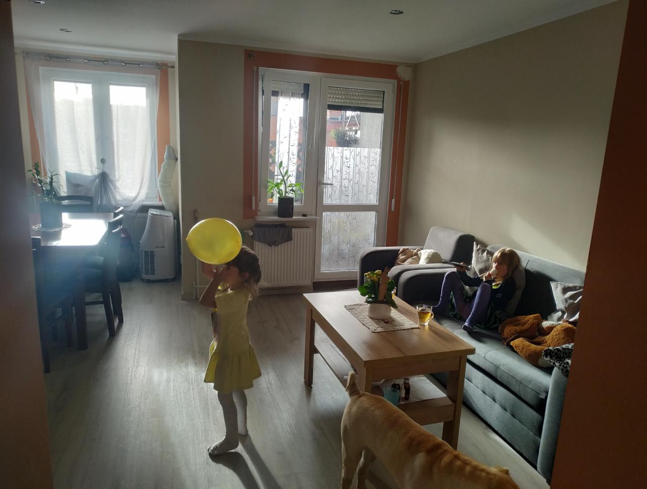 Dziewczynka w żółtej sukience dmucha żółty balon w jasnym pokoju. Druga dziewczynka odpoczywa na szarej sofie, oglądając telewizję. Pies w rogu kadru.