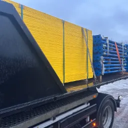 Ciężarówka z otwartą skrzynią ładunkową przewozi żółte płyty szalunkowe i niebieskie podpory budowlane zabezpieczone pasami transportowymi.