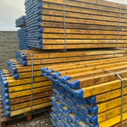 Stosy drewnianych dźwigarów budowlanych z niebieskimi zabezpieczeniami na końcach, spięte metalowymi taśmami, ułożone na paletach na placu budowy.