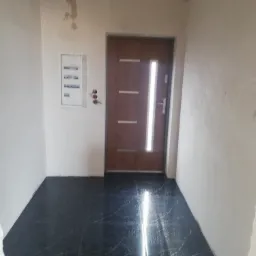 Korytarz w trakcie prac wykończeniowych: ciemna, błyszcząca posadzka z marmurkowym wzorem, surowe ściany z widocznymi śladami po zalaniu, brązowe drzwi z pionowym przeszkleniem i skrzynka...