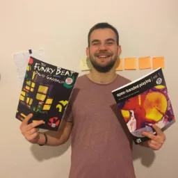 Uśmiechnięty mężczyzna prezentuje dwie książki do nauki gry na perkusji: 'The Funky Beat' i 'Open-Handed Playing Vol. 1'. W tle widoczne notatki i karteczki.