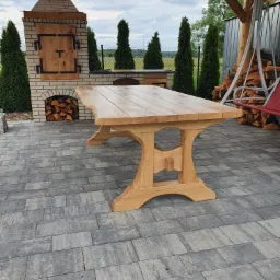 Solidny, drewniany stół z blatem o nieregularnych krawędziach, ustawiony na kostce brukowej w altanie z grillem z cegły i zapasem drewna w tle.