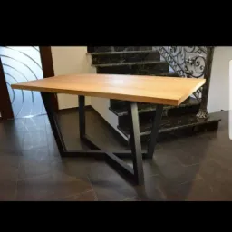 Stół z blatem z jasnego drewna i geometryczną, czarną podstawą metalową na tle ciemnej podłogi i schodów z metalową balustradą.