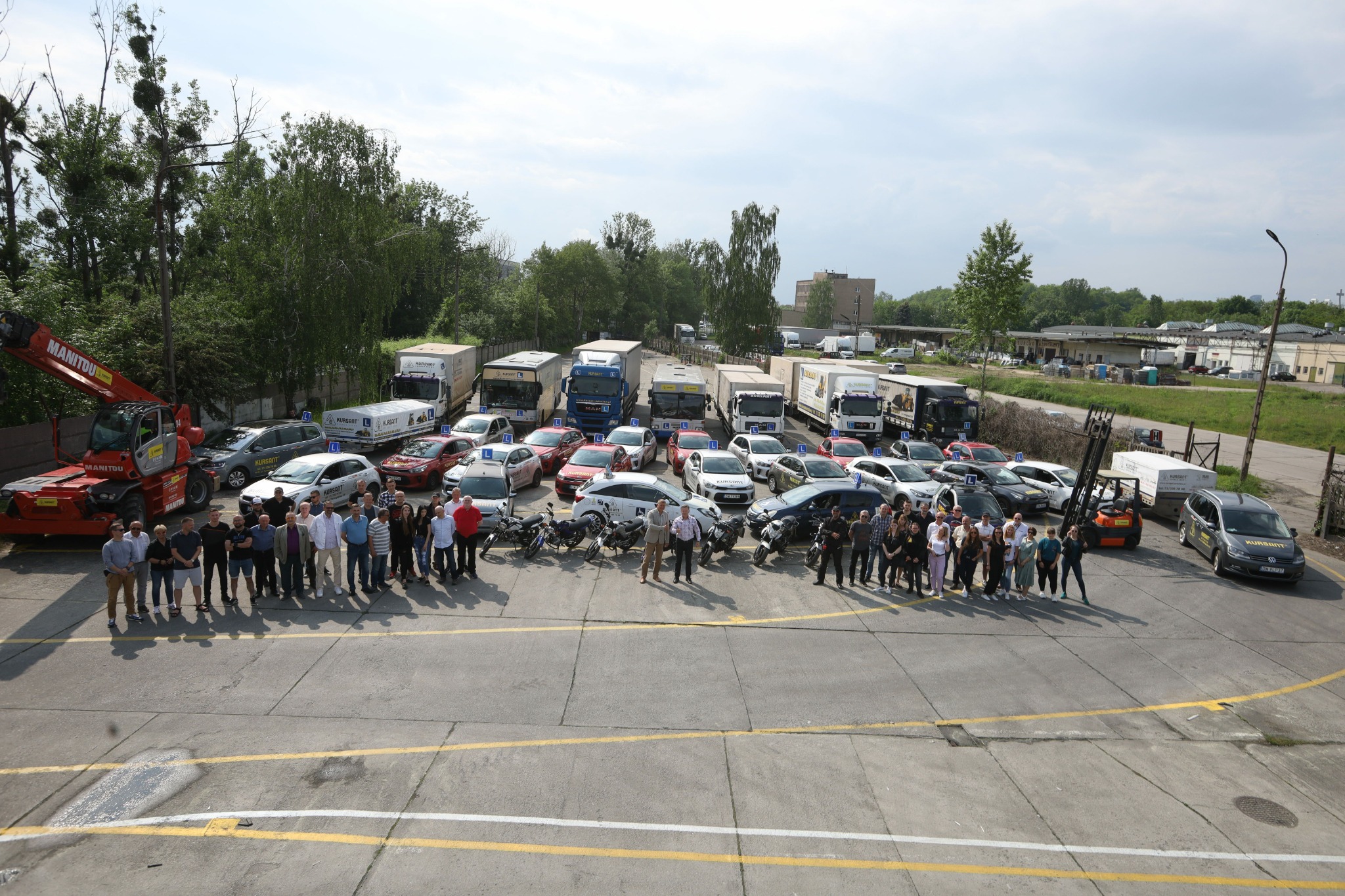 Grupa osób przed samochodami i motocyklami oznaczonymi jako pojazdy do nauki jazdy, na placu manewrowym. W tle drzewa i budynki.