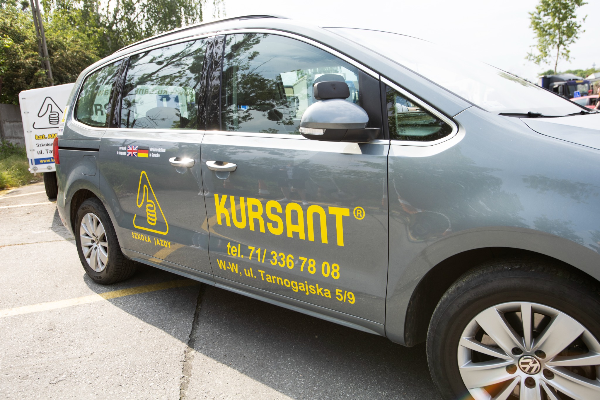 Szary samochód z żółtym napisem 'KURSANT' i logo Szkoły Jazdy, zaparkowany na parkingu. Widoczne numery telefonu i adres. Flagi UK i Niemiec na karoserii.