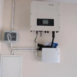 Biały inwerter SOFAR zamontowany na ścianie obok skrzynki elektrycznej z widocznymi przewodami i rurkami PCV, dodatkowo widoczne rękawice ochronne i dokumentacja techniczna.