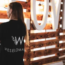 Kobieta w czarnej koszulce z logo 'Weselomania' na tle drewnianej ściany z napisem 'LOVE' i dekoracjami roślinnymi.
