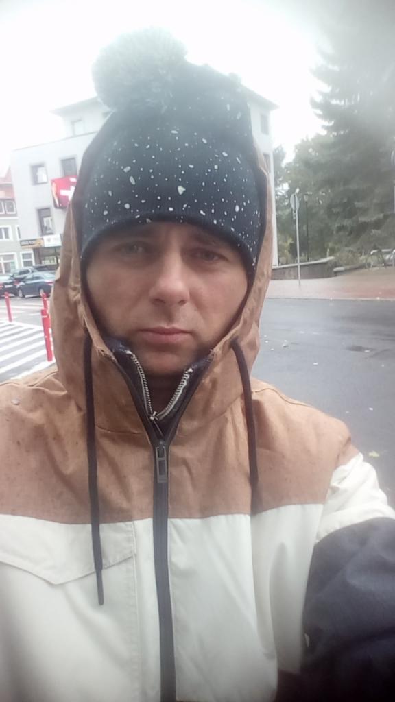 Portret mężczyzny w kurtce z kapturem i czapce z pomponem na tle miejskiego krajobrazu. Widoczne budynki, drzewa i fragment przejścia dla pieszych.