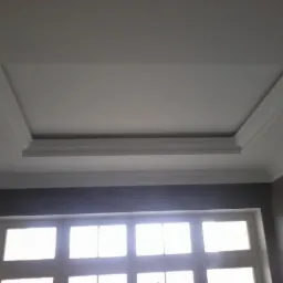 Dekoracyjny sufit podwieszany z białą sztukaterią, widok z dołu, szara ściana i okno z białymi ramami w tle.