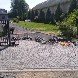 Roboty ziemne Minikoparka - Układanie kostki granitowej na podjeździe, widoczne rozłożone kamienie, narzędzia i częściowo ułożona nawierzchnia w kształcie wachlarza, brama wejściowa i zieleń w tle.