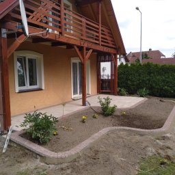 Roboty ziemne Minikoparka - Ułożona kostka brukowa tworząca obrzeże rabaty kwiatowej przed domem z drewnianym balkonem, fragment trawnika i rośliny wewnątrz rabaty.