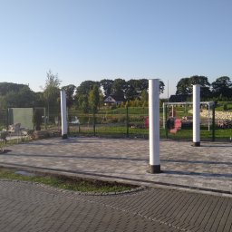 Roboty ziemne Minikoparka - Ułożona kostka brukowa w kolorze szarym z obramowaniem z ciemniejszej kostki, otoczona trawnikiem i białymi słupami, w tle widoczny ogród i drzewa.