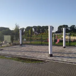 Ułożona kostka brukowa w kolorze szarym z obramowaniem z ciemniejszej kostki, otoczona trawnikiem i białymi słupami, w tle widoczny ogród i drzewa.