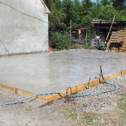 Świeżo wylany fundament z drewnianym szalunkiem, widoczny niebieski wąż doprowadzający wodę i pies rasy owczarek niemiecki stojący na skraju placu budowy.