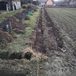 Wykopane rowy w ziemi, prawdopodobnie pod instalacje, widoczne sznurki wyznaczające linie, narzędzie pracy leżące obok rowu, przygotowane do zasypania ziemią. W tle zabudowania.