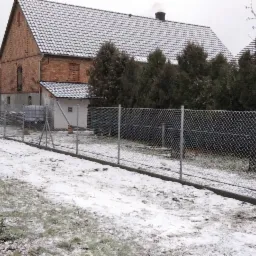 Nowo postawione ogrodzenie z siatki na posesji w zimowej scenerii, widoczny budynek mieszkalny w tle, częściowo pokryty śniegiem.