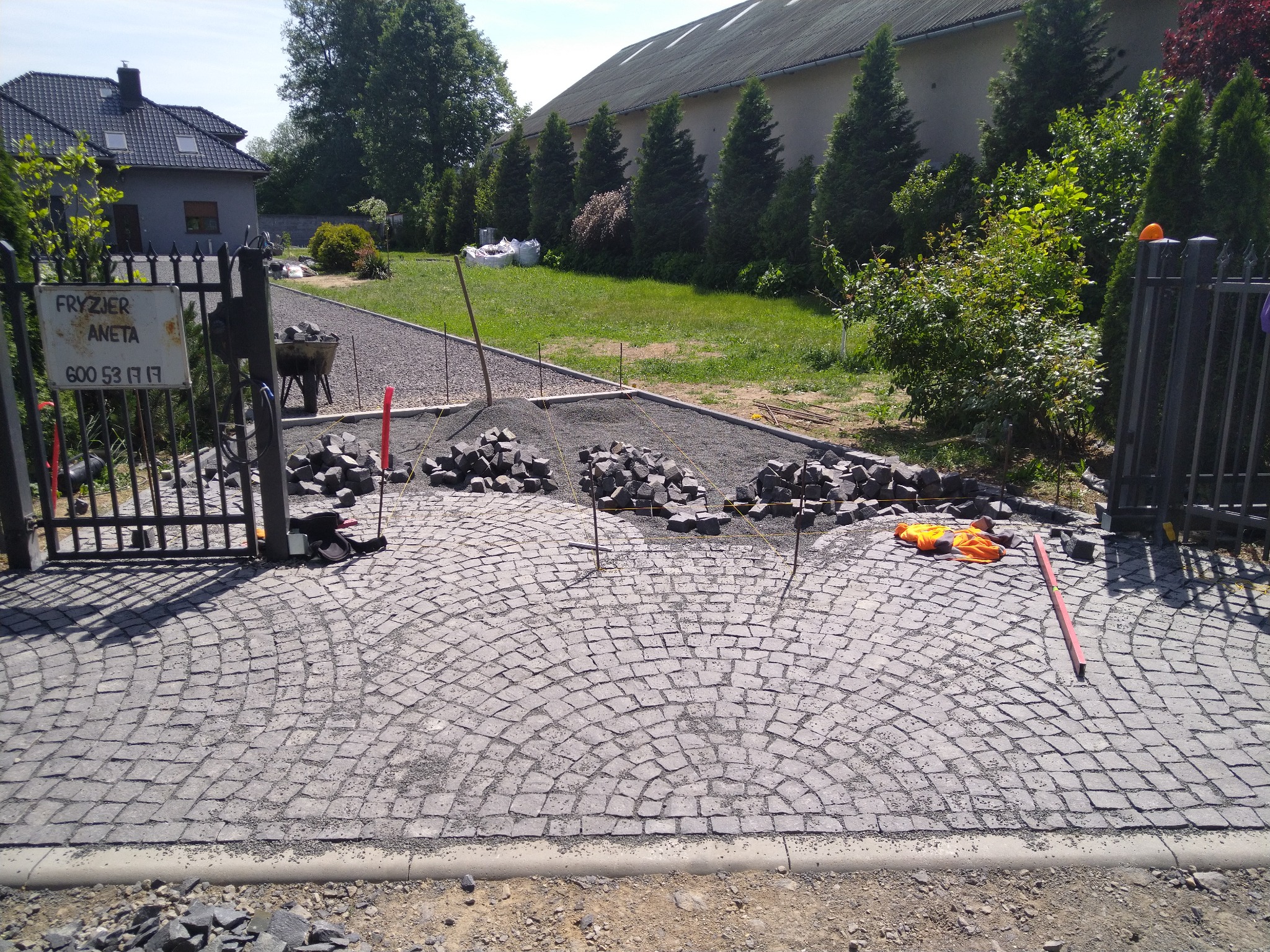 Układanie kostki granitowej na podjeździe, widoczne rozłożone kamienie, narzędzia i częściowo ułożona nawierzchnia w kształcie wachlarza, brama wejściowa i zieleń w tle.