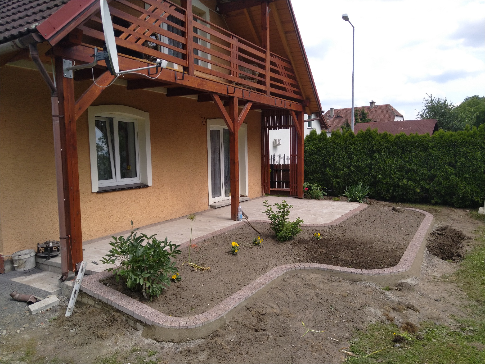 Ułożona kostka brukowa tworząca obrzeże rabaty kwiatowej przed domem z drewnianym balkonem, fragment trawnika i rośliny wewnątrz rabaty.