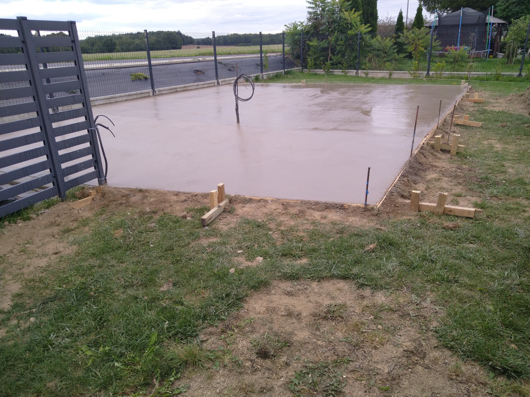 Świeżo wylany fundament pod budowę w Wykrotach, z widocznymi deskami szalunkowymi i wystającymi przewodami instalacyjnymi, otoczony zielonym trawnikiem i ogrodzeniem panelowym.