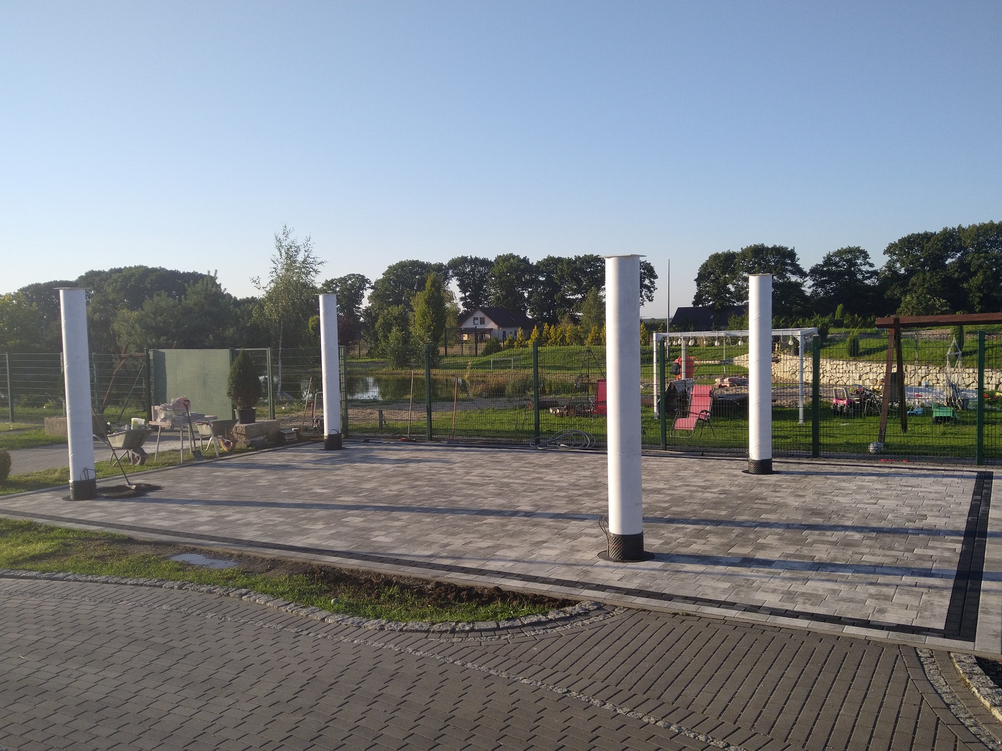 Ułożona kostka brukowa w kolorze szarym z obramowaniem z ciemniejszej kostki, otoczona trawnikiem i białymi słupami, w tle widoczny ogród i drzewa.