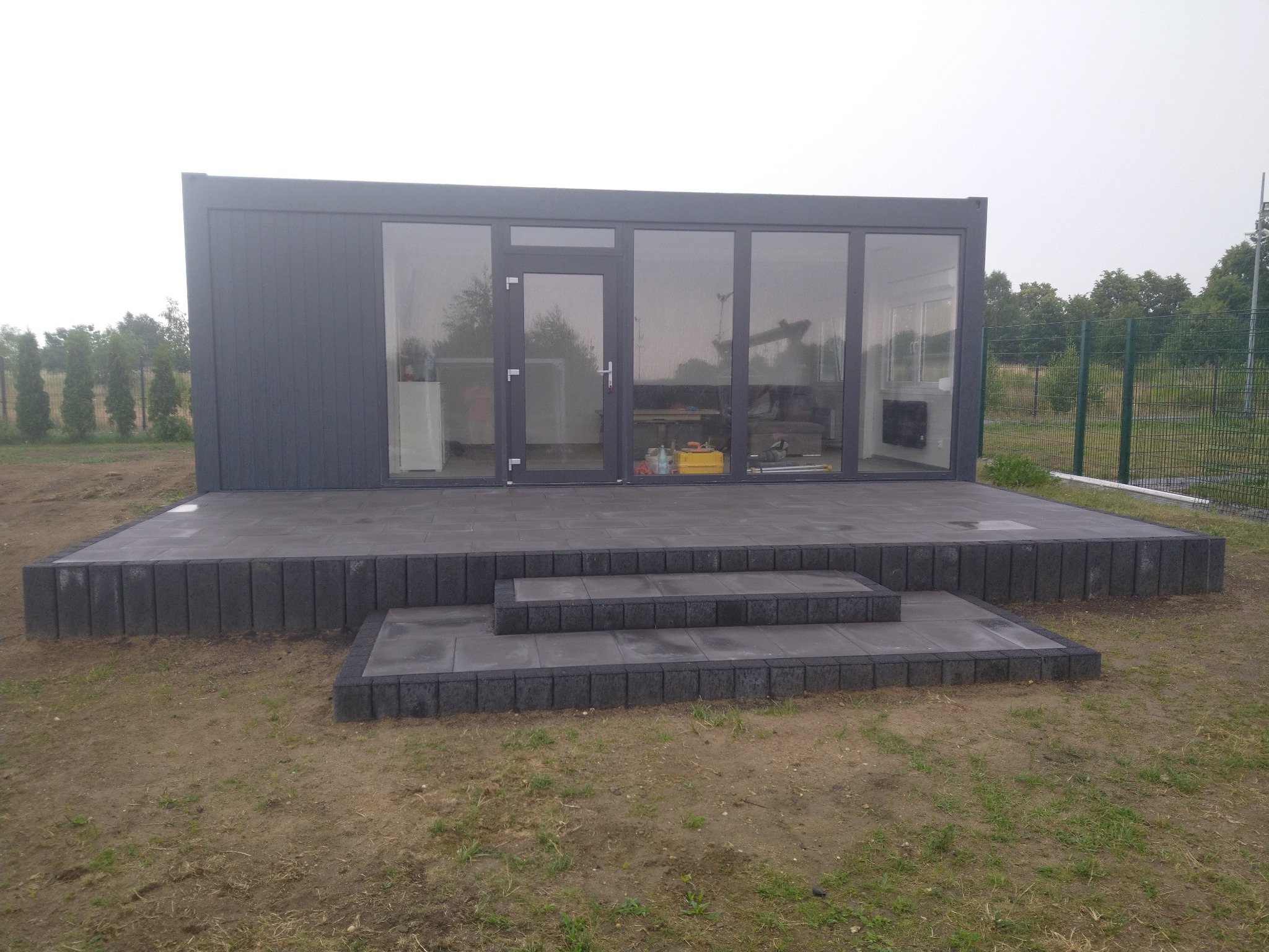 Nowoczesny, szary pawilon z dużymi oknami i drzwiami, posadowiony na podwyższonym tarasie z kostki brukowej, z trzema schodkami prowadzącymi na trawnik.
