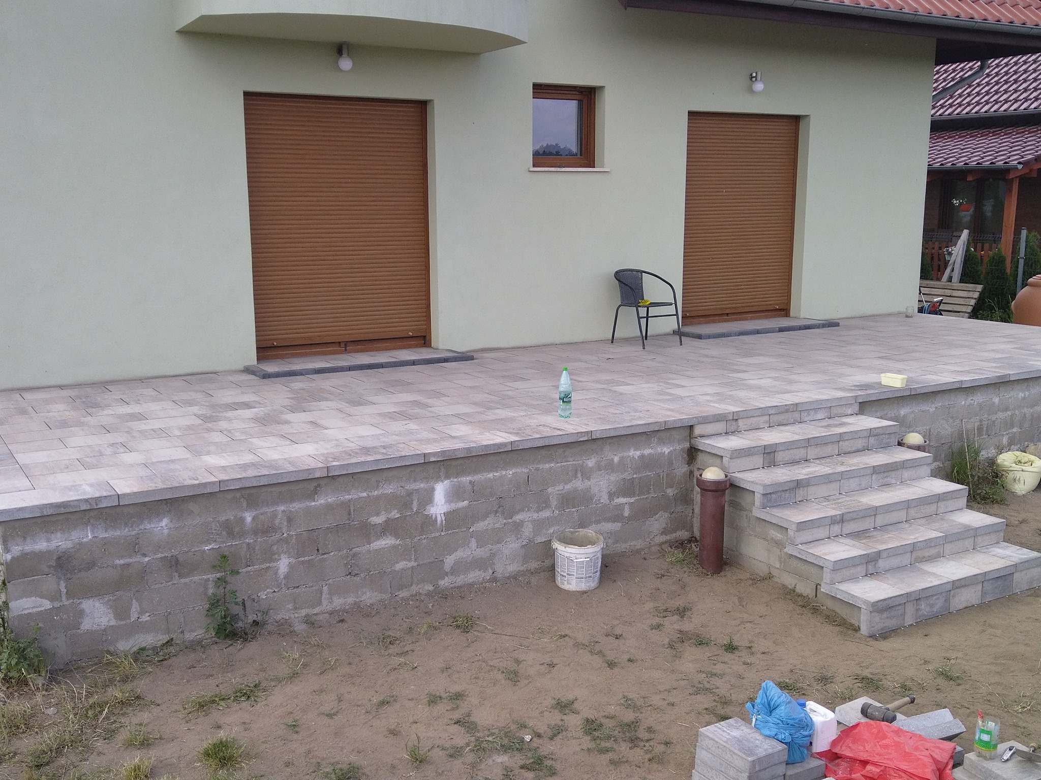 Wykończony taras z kostki brukowej przed budynkiem z brązowymi roletami, widoczne schody z kostki, krzesło i butelka wody na tarasie, narzędzia i resztki materiałów budowlanych na ziemi.