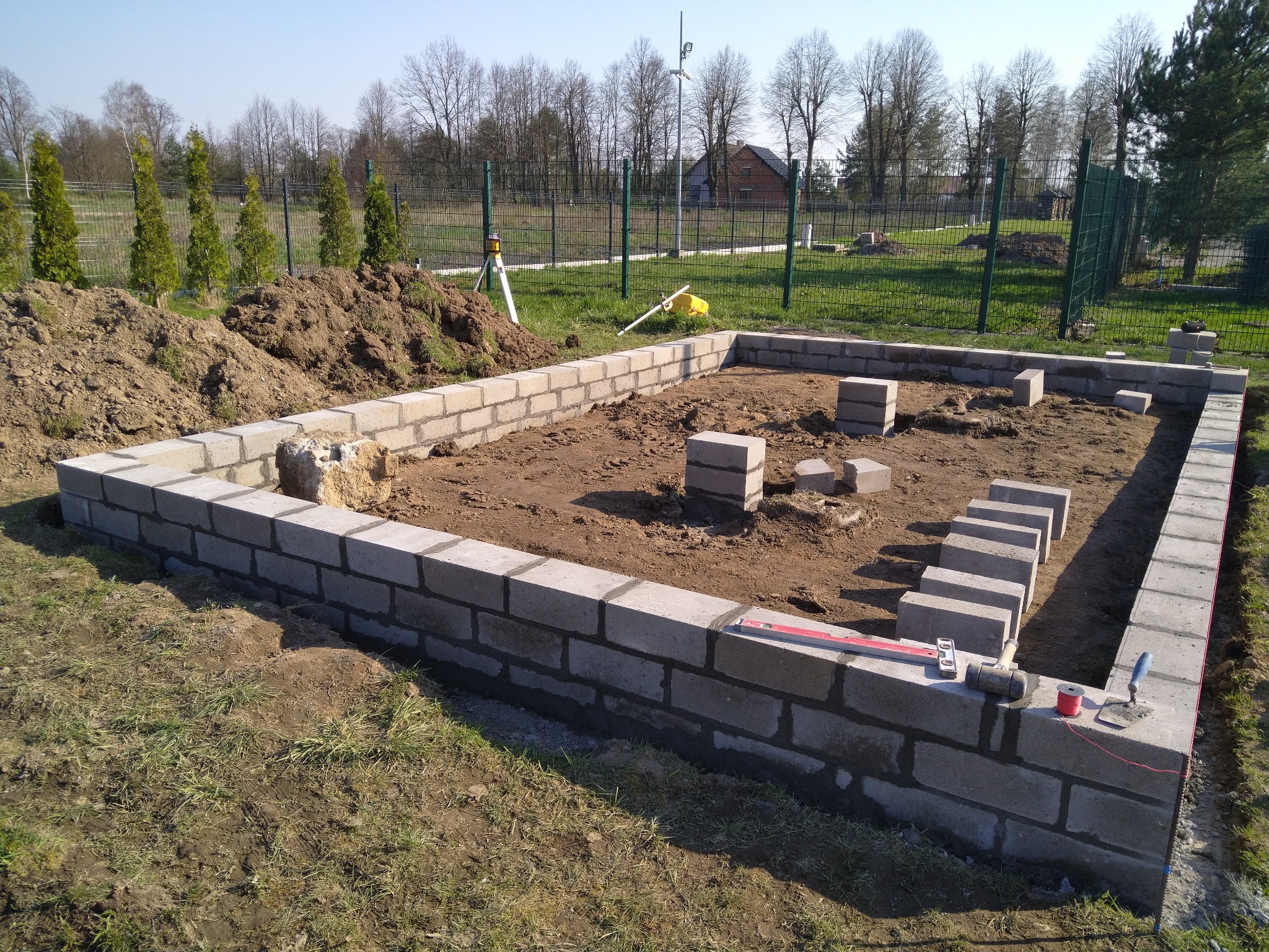 Wznoszenie fundamentów z bloczków betonowych, widoczne narzędzia murarskie, poziomica, sznurek traserski, oraz laserowy niwelator w tle, ziemia wokół.