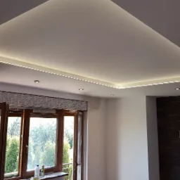 Nowoczesny salon z podwieszanym sufitem z oświetleniem LED, widok na okno z roletami i dekoracyjną ścianę z kamienia.