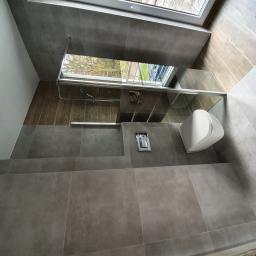 Usługi remontowe - Nowoczesna łazienka z szarymi płytkami, kabiną prysznicową z widokiem na zewnątrz przez okno dachowe, wiszącą toaletą i ukrytą spłuczką.