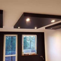 Usługi remontowe - Nowoczesny salon z czarnymi podwieszanymi panelami sufitowymi z punktowym oświetleniem LED, kontrastującymi z białymi ścianami i czarną ścianą z oknem.